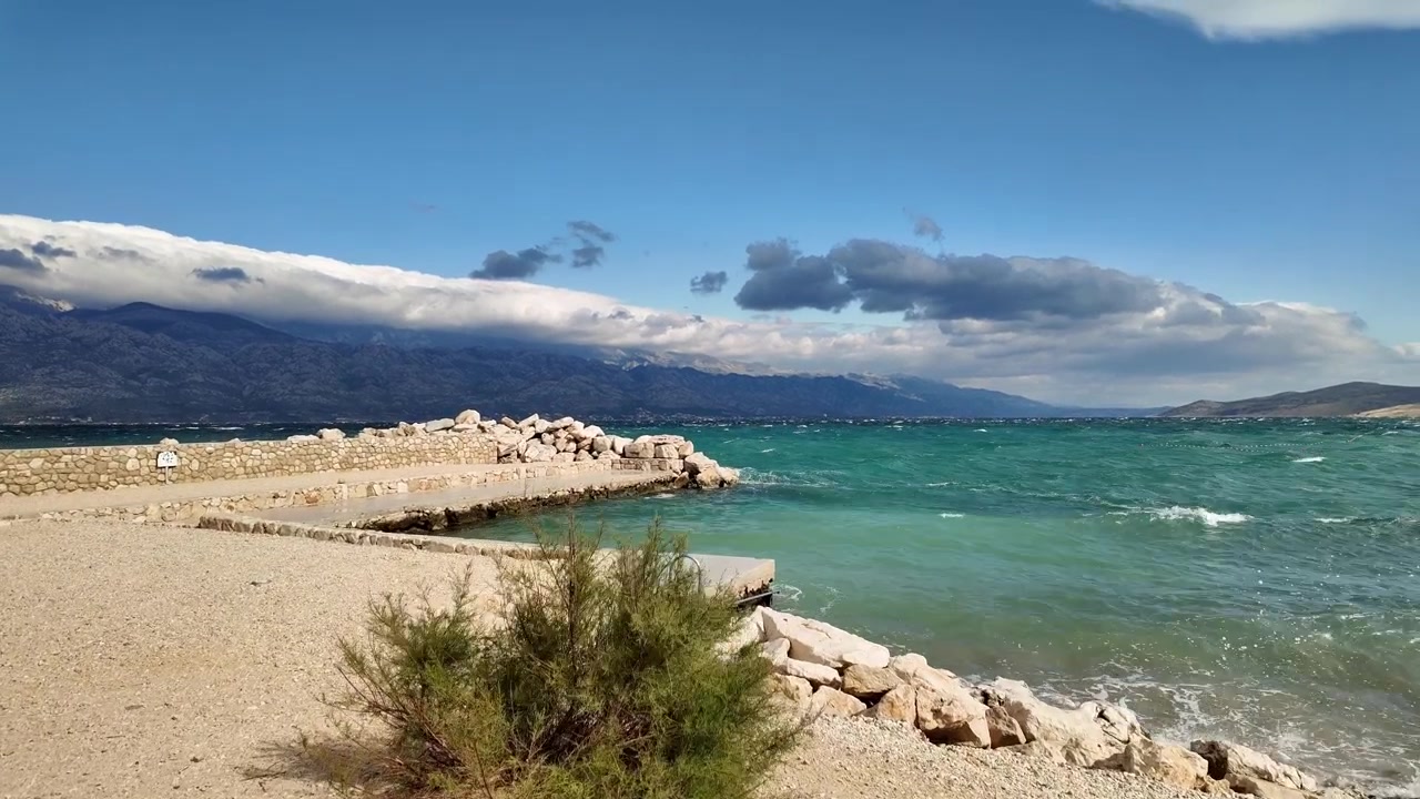 Razanac'ın boş plajında şiddetli Bura rüzgarında yürüyüş, dalgalar çarpıyor, bir çalılık şiddetle sallanıyor, taş duvar koruması ve uzakta Velebit görünüyor.