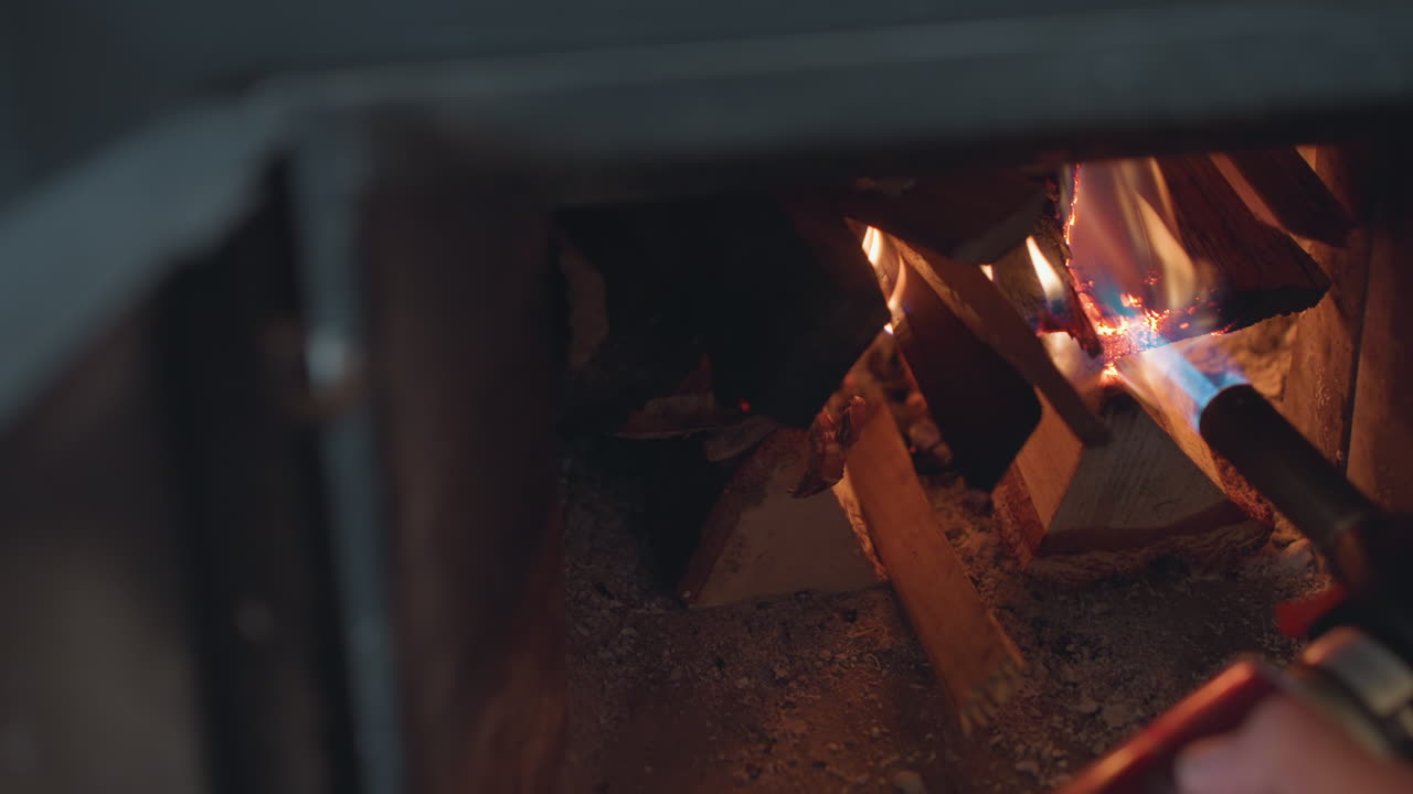 Extreme close up of person using blue flame torch to ignite stacked firewood inside dark fireplace, flame catches gradually as wood begins to burn, glowing with soft embers in warm indoor environment