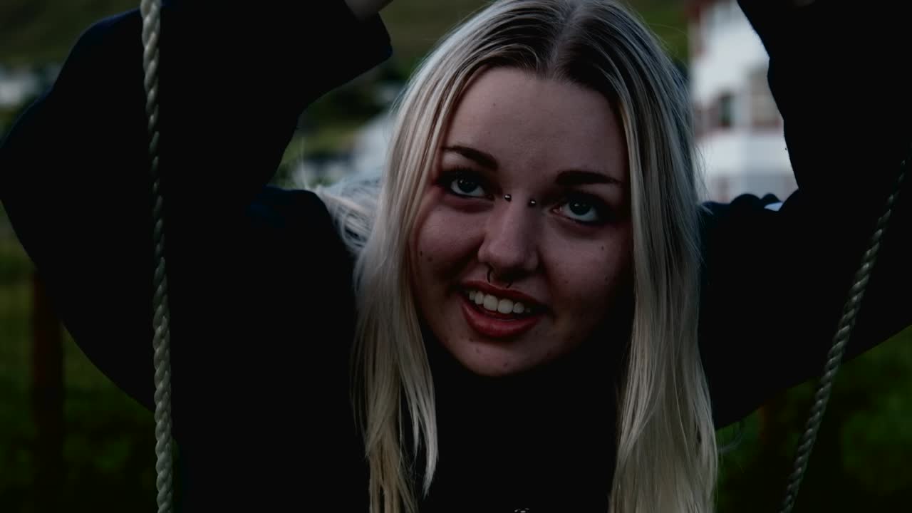 счастливая улыбающаяся молодая блондинка сидит на качелях в холодный зеленый весенний вечер