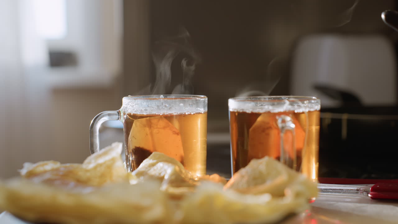 Primo piano di due tazze di vetro fumanti riempite con tisana calda e bustine di tè all'interno, vapore che sale dalla superficie, pancake serviti su un piatto in primo piano, accogliente ambientazione per la colazione