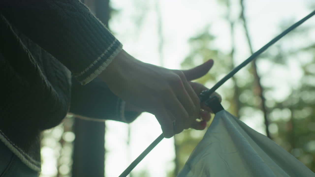 Close up of person in knitted sweater preparing tent by hooking pole through fabric loop in forest with soft natural lighting and bokeh background