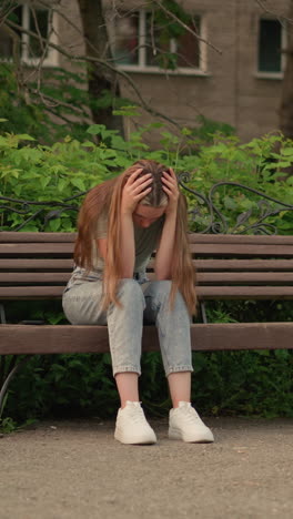 una mujer se sienta en un banco de madera, en una postura sombría, sus largos cabellos caen en cascada mientras apoya su cabeza con las manos, rodeada de exuberante vegetación, árboles y un edificio urbano en el fondo.