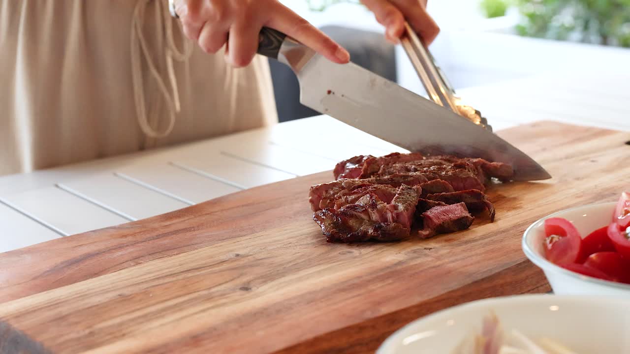 A person slices beef steak on a wooden board in a bright kitchen. Natural lighting enhances the fresh, culinary atmosphere