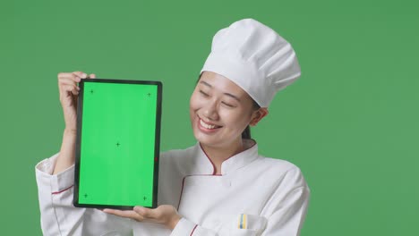 close up of asian woman chef smiling and showing green screen tablet to the camera while standing in the green screen background studio