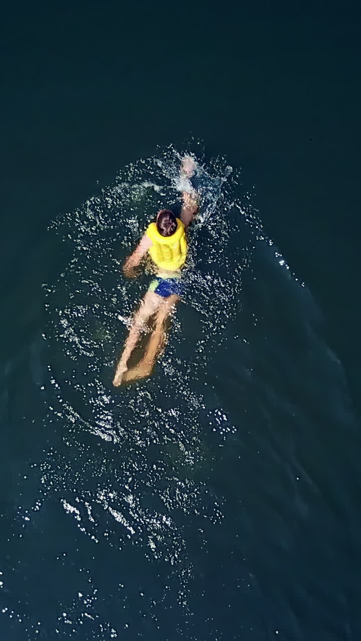 Aerial shot of a boy swimming in clear sea water. Top view of a boy swimming away camera in the middle of the ocean Vertical video