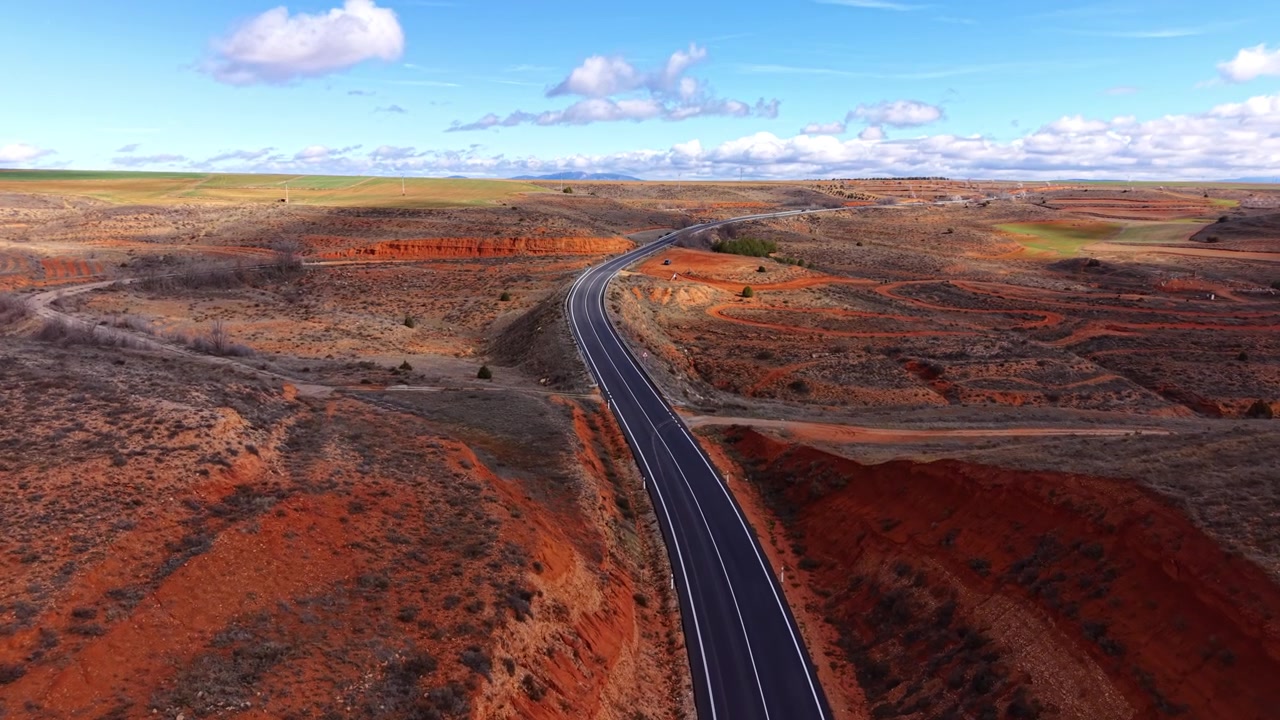 Gewundene Asphaltstraße, die sich durch leuchtend rotes Felsgelände unter einem teilweise bewölkten blauen Himmel schlängelt und sich in Richtung ferner grüner Ebenen und Berge am Horizont erstreckt