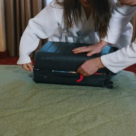 couple in bathrobes closing suitcase together. husband helping his wife to close travel bag at the end of their vacation in hotel. close-up