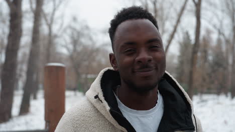 smiling man wearing winter jacket standing outdoors in snowy environment with bare trees in background, his warm expression contrasts with the cold atmosphere