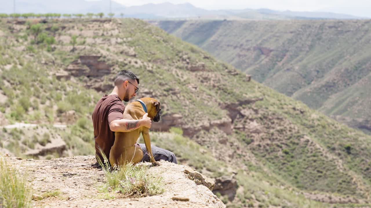 Man hugging Boxer dog while sitting on cliffside in green valley