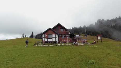 la panorámica a la derecha muestra una cabaña de madera en un verde prado de montaña con algo de niebla en el fondo