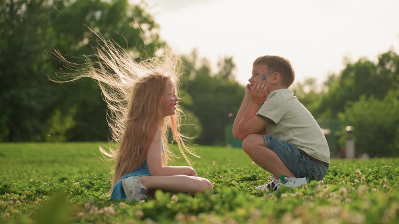 Toddlers seated on grass while parents sway fabric over them playfully, warm light filtering through trees, casual summer setting, family interaction showing gentle care and joyful connection