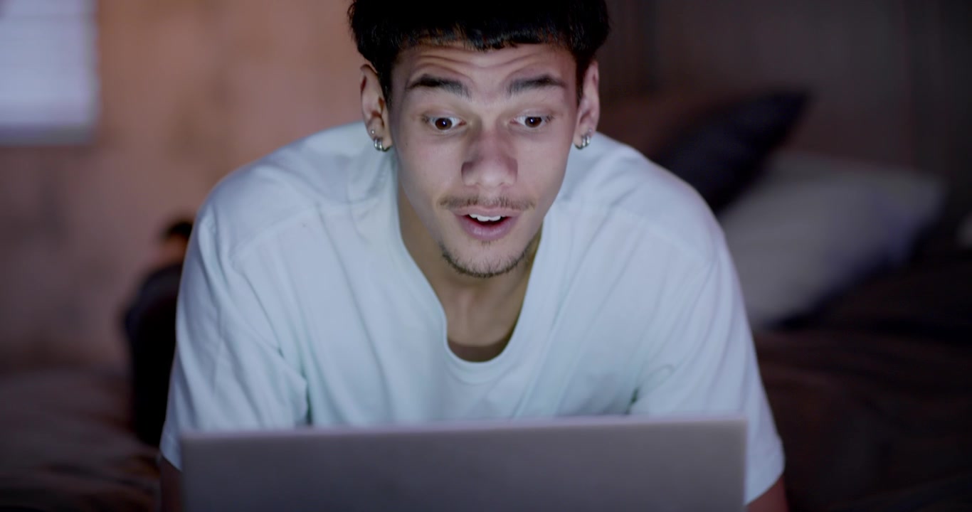 African American man watching laptop on bed, lifting head as logo fills frame, reacting to clip