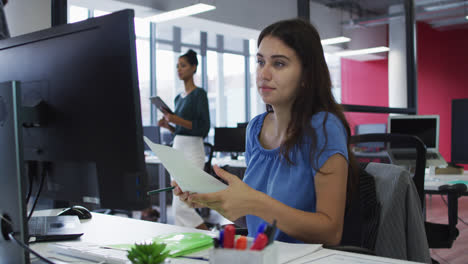 mujer de negocios caucásica sentada en un escritorio usando una computadora y mirando un documento en una oficina ocupada