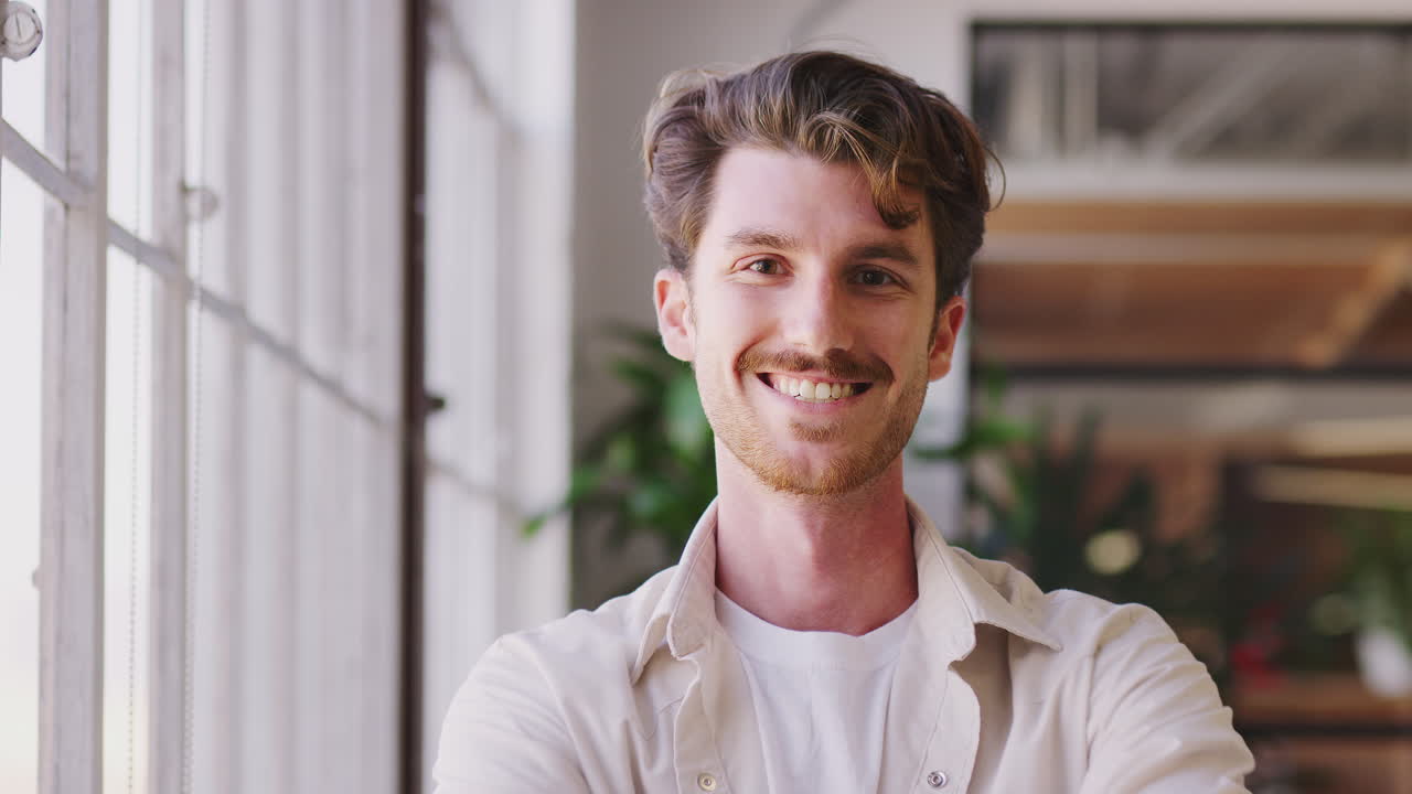 밀레니엄 백인 남성 크리에이티브가 사무실에 서서, 카메라로 돌아서 웃으며, 클로즈업