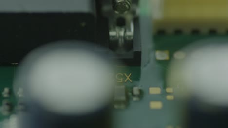 macro view of a green circuit board featuring various components, focus on a small black chip, with other elements like a rectangular component, a blue capacitor, and silver oscillator also visible