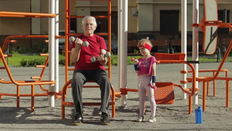 Granddaughter-and-grandfather-doing-fitness-exercises-with-dumbbells.-Senior-man-with-child-kid-girl