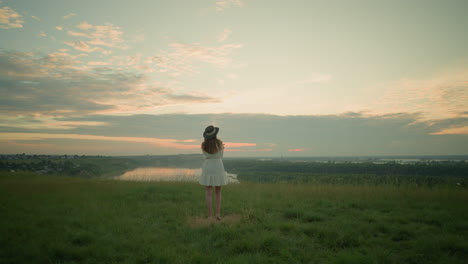 a woman in a white dress and hat stands thoughtfully in a grassy field overlooking a tranquil lake at sunset. she gazes at the serene environment, capturing a peaceful and reflective moment