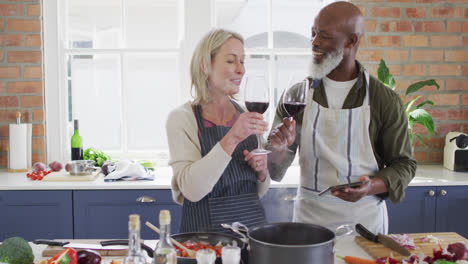 Pareja-De-Ancianos-De-Raza-Mixta-Usando-Delantales-Bebiendo-Vino-Mientras-Cocina-En-La-Cocina-De-Casa