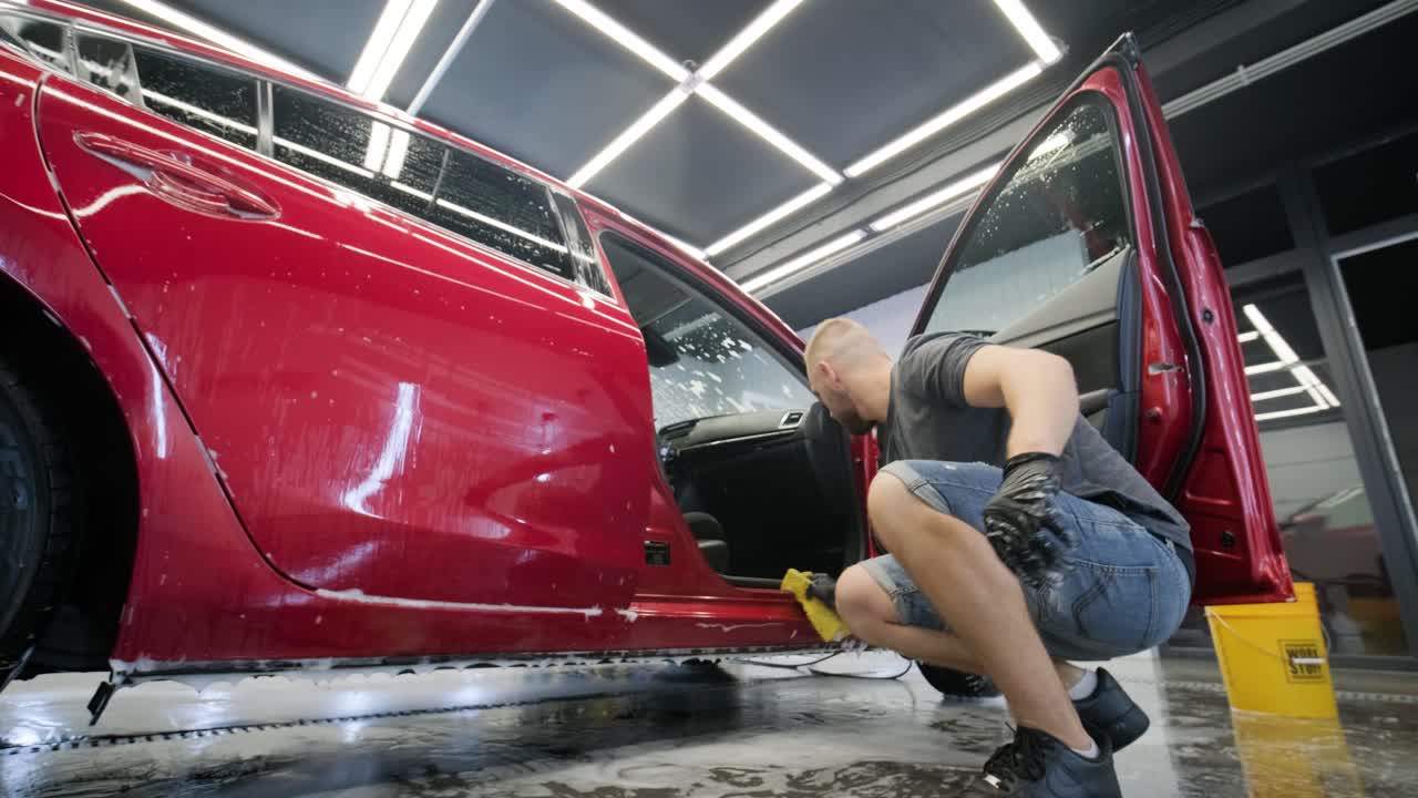 un trabajador lavando un coche rojo en un lavado de coches con un paño amarillo.