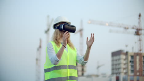 una mujer con gafas de realidad virtual, casco y chaleco en el fondo de la construcción controla las manos de la interfaz y comprueba la calidad de la construcción y el desarrollo del proyecto y el plan de desarrollo y el paisaje.