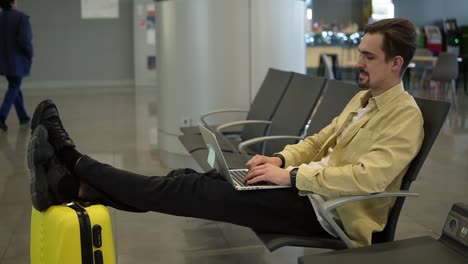 Un-Joven-Aburrido-Está-Sentado-En-El-Aeropuerto,-Esperando-Que-Salga-El-Avión-Y-Usando-Una-Computadora-Portátil.-Escribiendo-En-La-Computadora-Portátil,-Sosteniendo-Las-Piernas