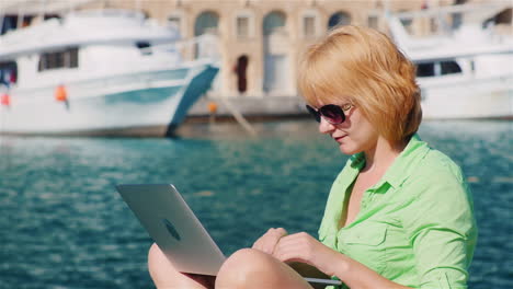 Girl-Tourist-Uses-A-Laptop-On-The-Background-Of-The-Urban-Landscape-With-Yachts