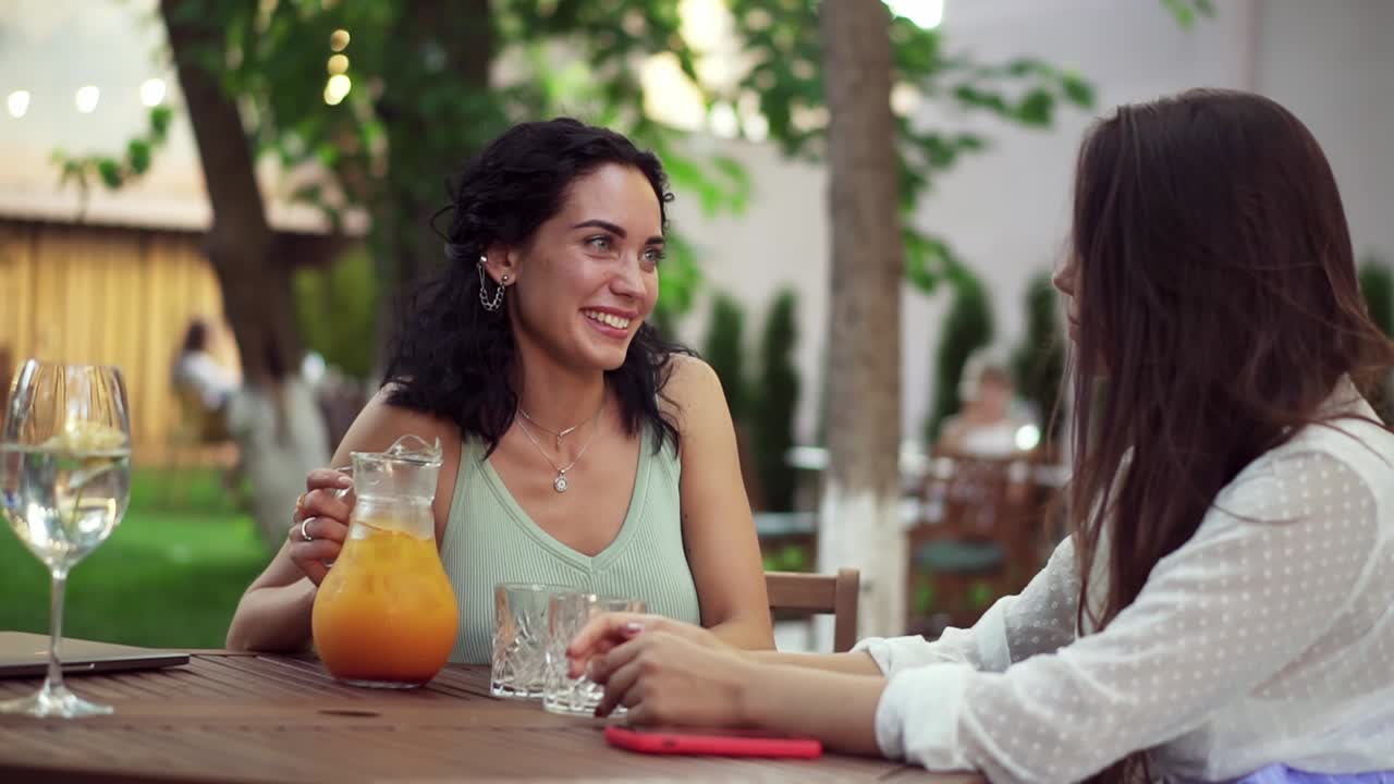 personas, comunicación y concepto de amistad - mujeres jóvenes sonrientes bebiendo jugo de naranja y hablando en una cafetería al aire libre en verano