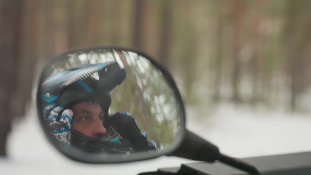 Pilota con casco riflesso nello specchio, visiera e guanto fantasia visibili, sfondo di bosco di pini innevato, primo piano di espressione contemplativa, pilota che scruta il sentiero pronto per un'avventura fuoristrada estrema.