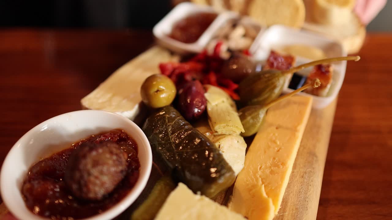 Camera smoothly pans over assorted cheeses, meats, olives, and dips on rustic wooden board