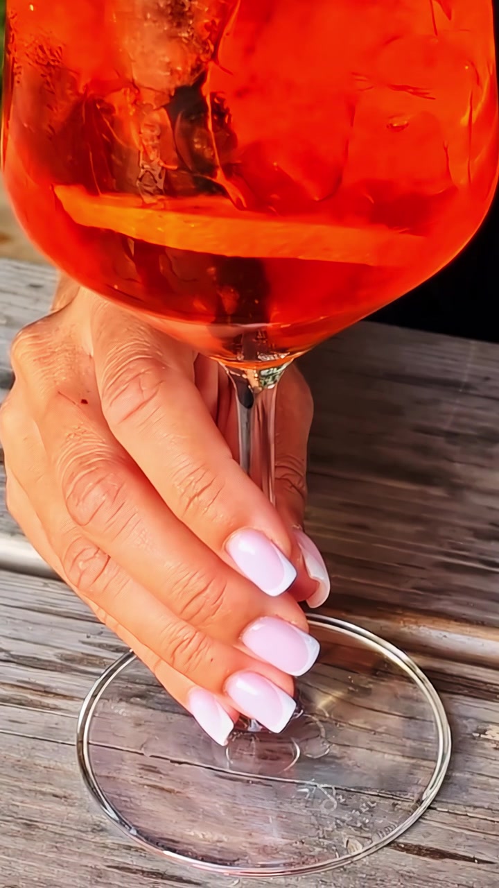 Genç bir kadının bakımlı eli, ahşap bir masada serinletici bir Aperol Spritz bardağı tutuyor.