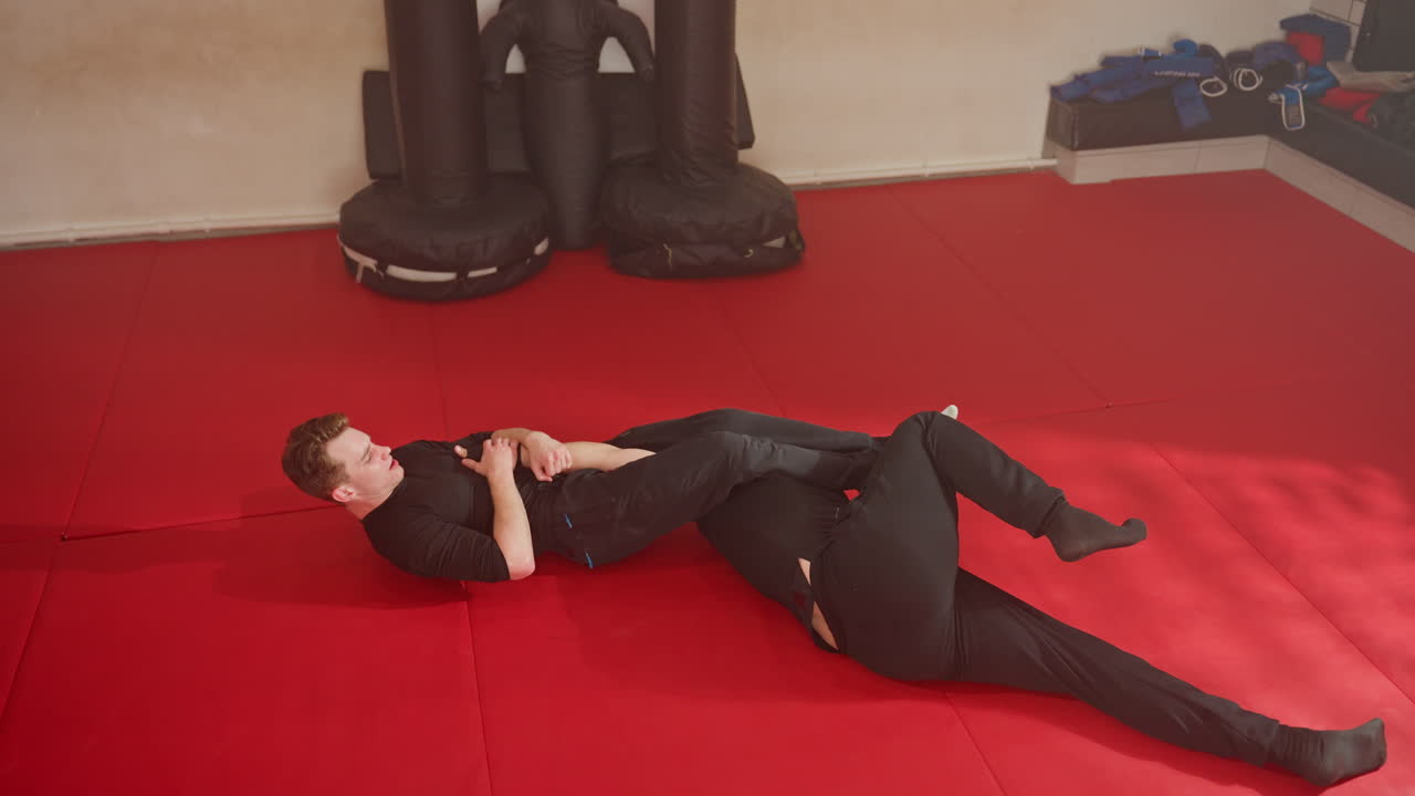 Wrestlers engage in intense grappling on red mat during training session, one athlete applying hold from behind while opponent struggles to break free, physical endurance in combat practice