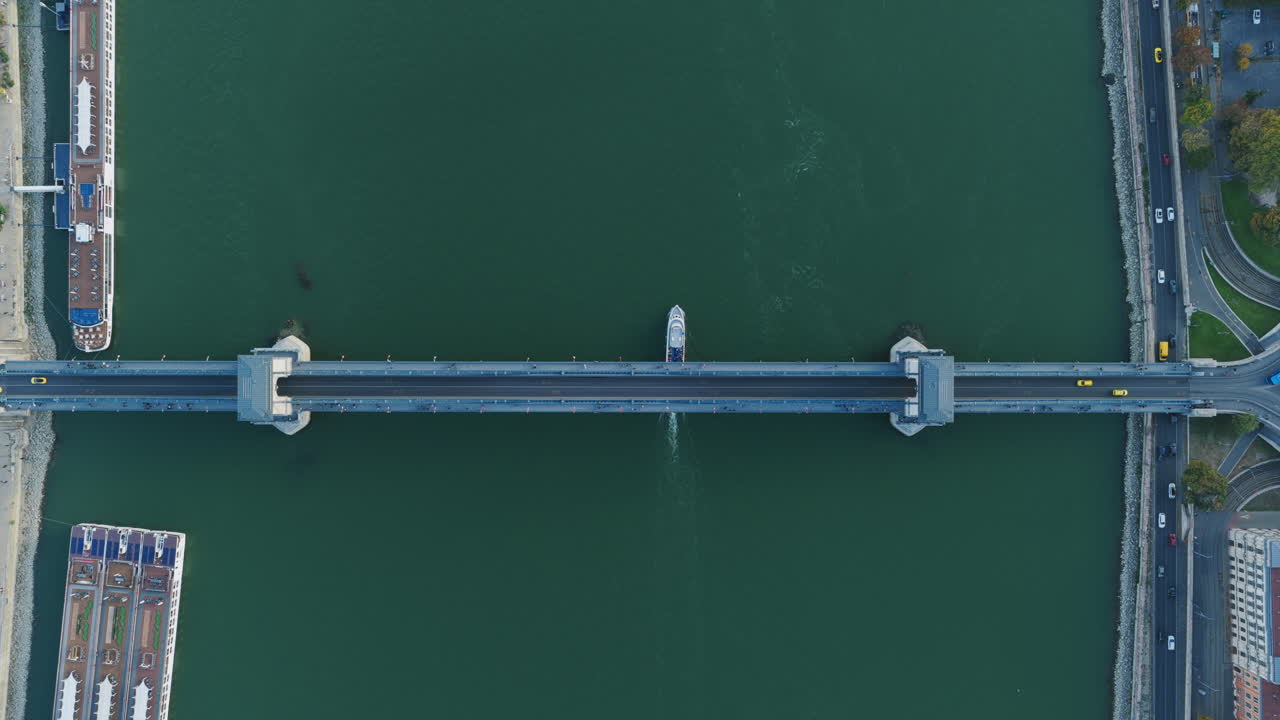 Perfectly symmetrical top-down shot of a ferry gliding under a Budapest bridge over the Danube River