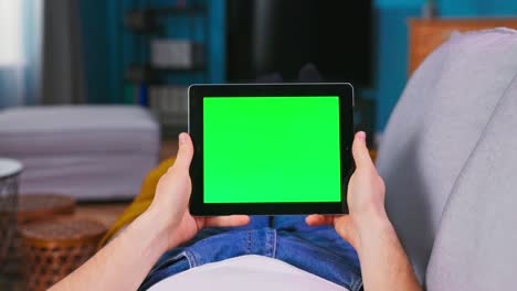 hombre joven en casa acostado en un sofá usando con pantalla de maqueta verde tableta computadora en modo paisaje. niño está navegando por internet, viendo contenido, videos, blogs