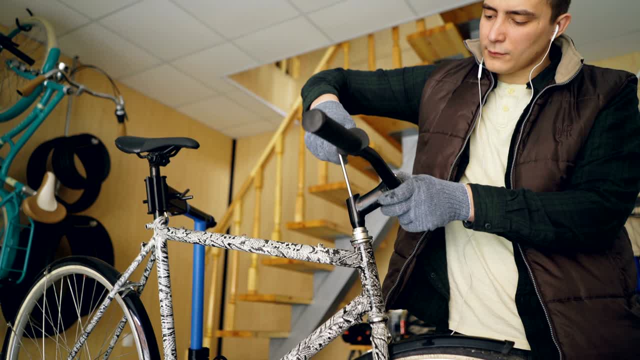 집중된 남성 기계공은 자전거 손잡이를 고치면서 이어폰으로 음악을 듣고, 열로 프레임에서 을 풀고, 유지보수 및 사람 개념을 확인합니다.