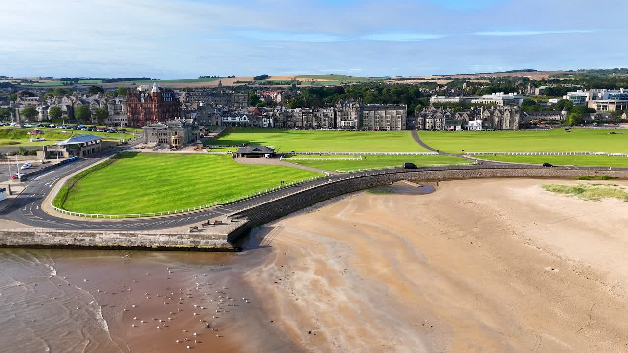 Aerial camera slowly pans across a Victorian brick building, lush golf course, and sandy beach under bright daylight in St Andrews, Scotland