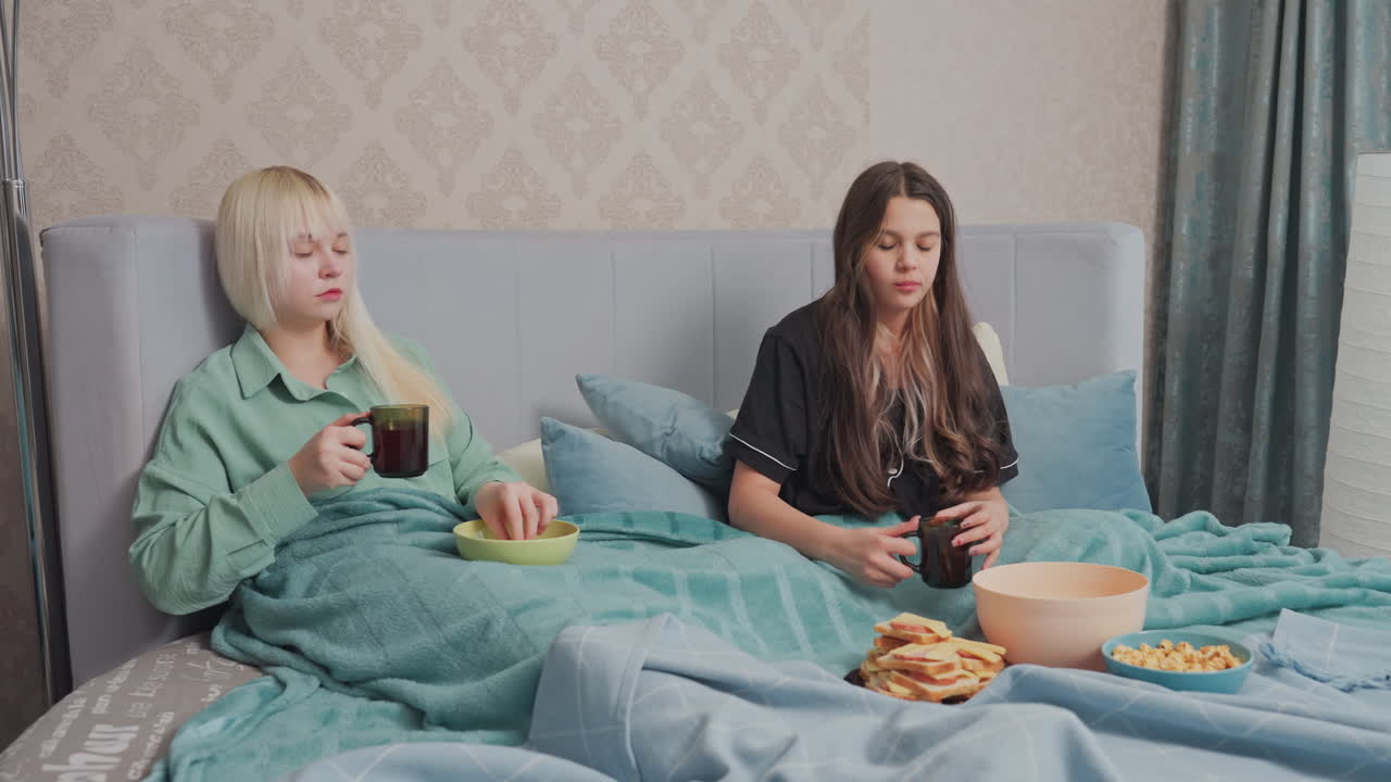 Two college girls relax in bed under teal blankets, enjoying a cozy snack moment, one girl in black pajamas hands over bowl of gummy candy while the other, holding a mug, smiles and receives it