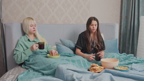 two college girls relax in bed under teal blankets, enjoying a cozy snack moment, one girl in black pajamas hands over bowl of gummy candy while the other, holding a mug, smiles and receives it