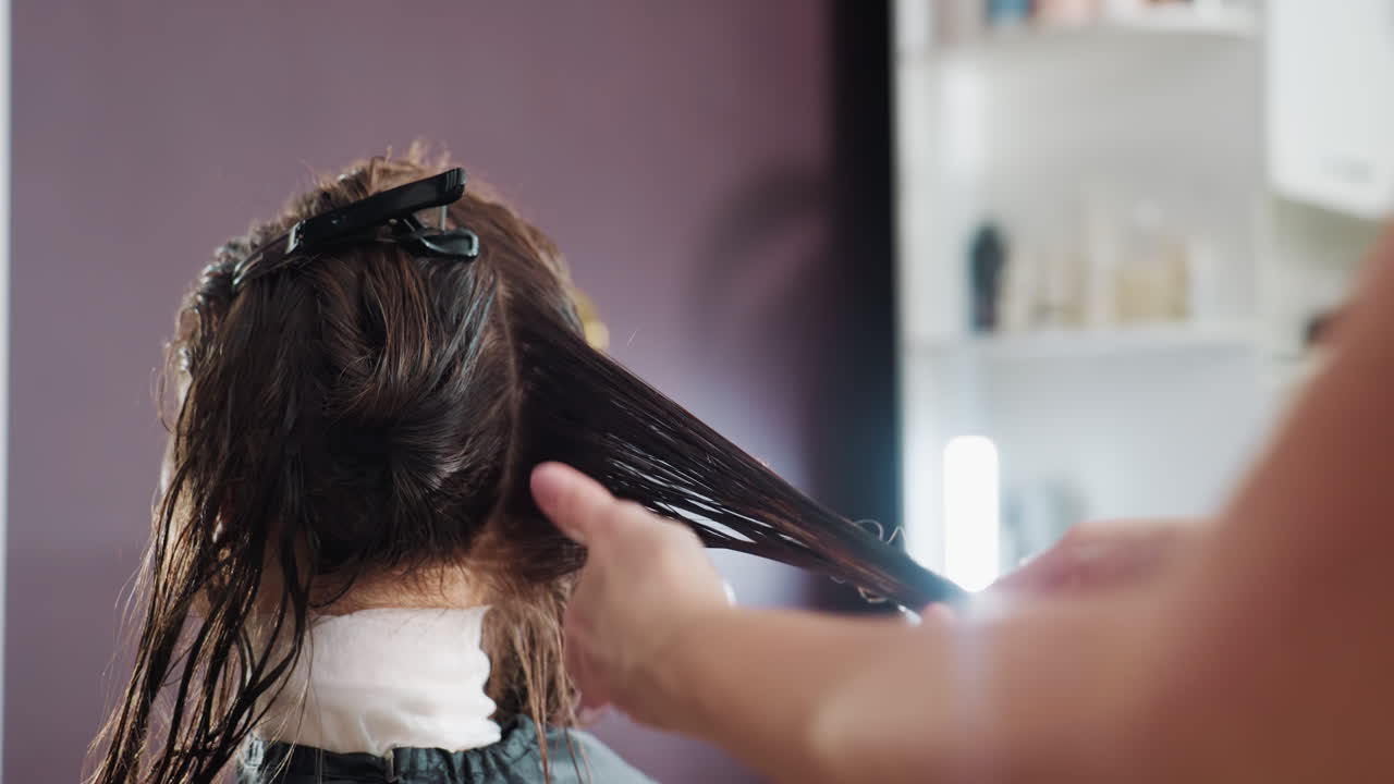 젖은 머리 뒷부분을 나누어 자르는 스타일리스트의 뒷모습, 클립과 가위로 머리를 잡고 있는 정확한 손길, 제품 선반과 집중된 기술이 돋보이는 친근한 분위기의 미용실 내부