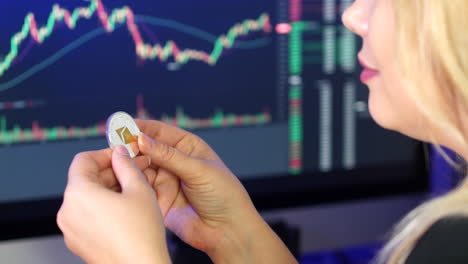 blonde woman holding and looking at ethereum coin in her hands in front of cryptocurrency trading graphs