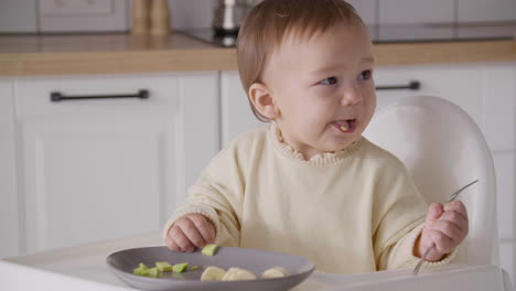 Cute-Baby-Girl-Eating-Avocado-Slices-Sitting-In-Her-High-Chair-In-The-Kitchen