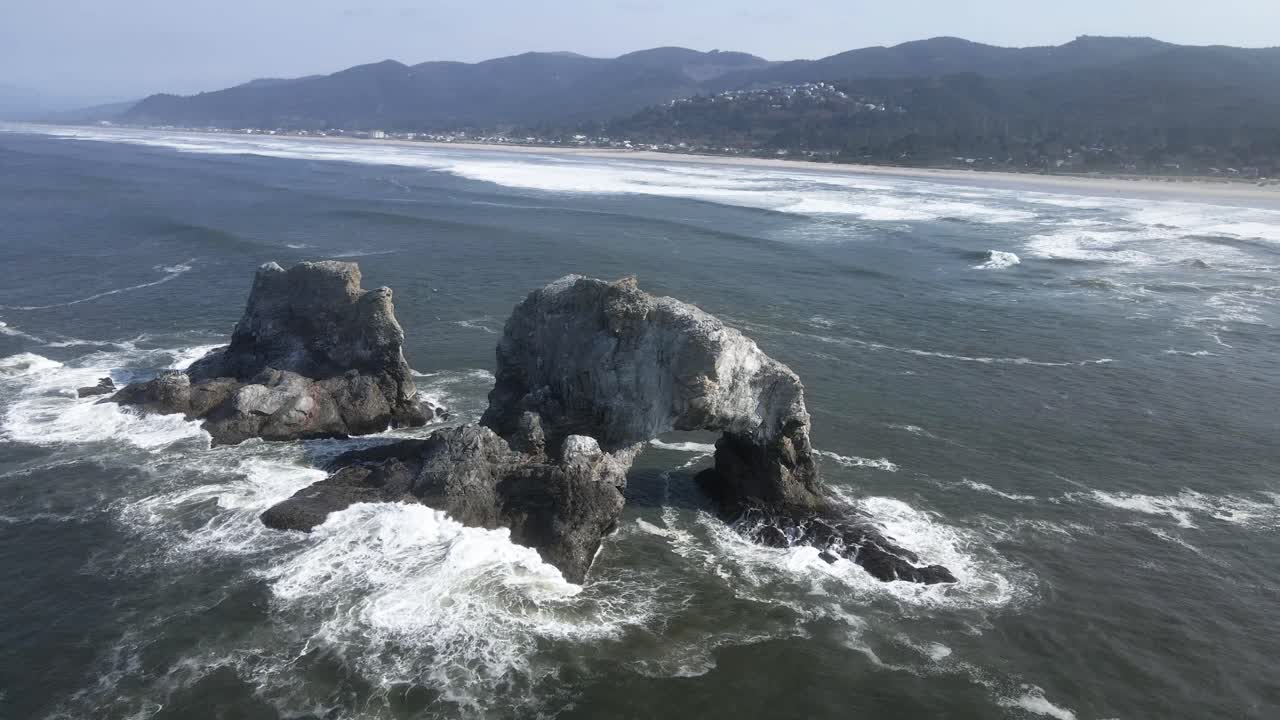 오레곤주 로커웨이 해변의 해안에서 떨어져 있는 쌍둥이 바위의 뒷면에 파도가 충돌합니다.