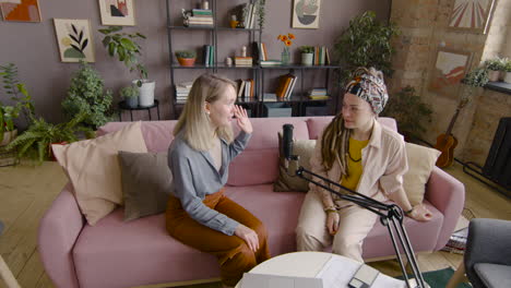 zoom out and top view of two women recording a podcast talking into a microphone while sitting on sofa in front of table with laptop and documents