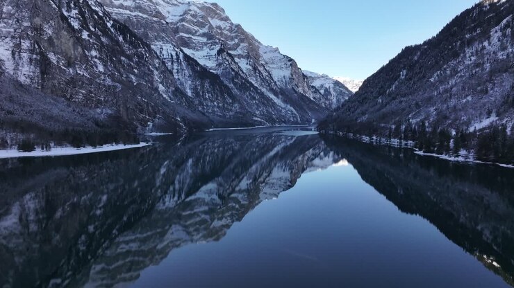 Breathtaking drone pushes towards aerial view of Klöntalersee, nestled in Klöntal, Glarus with snowy Swiss alps reflected in calm lake waters during serene winter.
