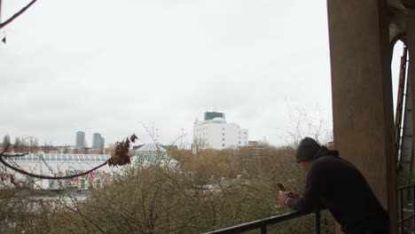 man walks to old abandoned building railing, stops to text send picture looking at cityscape