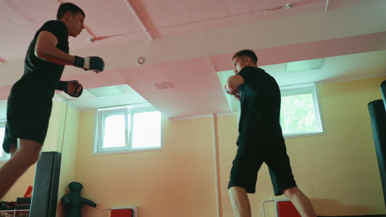 Two fighters sparring in gym on red mat, barefoot, wearing gloves, practicing martial arts with fast movements, focusing on punches, defense, strength combat training under natural window light