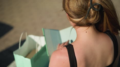 vista trasera de una mujer sentada en un banco al aire libre con una mancha en la espalda, recuperando un libro de una bolsa verde menta, abre el libro y hojea sus páginas mientras las sombras se proyectan en el suelo