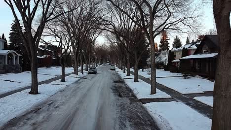 aerial-pan-out-sunset-twilight-drone-camera-flight-from-quiet-luxury-winter-residential-neighborhood-with-tall-old-bare-trees-pines-fur-parked-cars-on-the-snow-covered-roads-and-some-empty-drive-ways