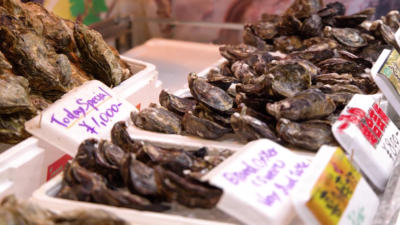 Close-up shot of fresh oysters in shells at a market stall with vendor hands visible.