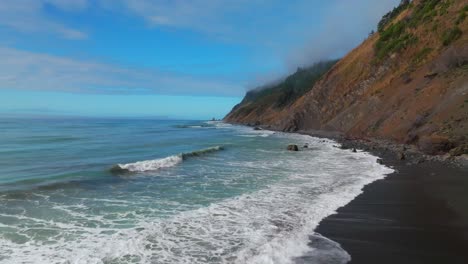 waves crashing surf black sand rugged coastline sunny morning blue sky mist fog layer usal beach campground lost coast trail california aerial drone pnw boulders rocks mountainside backwards motion