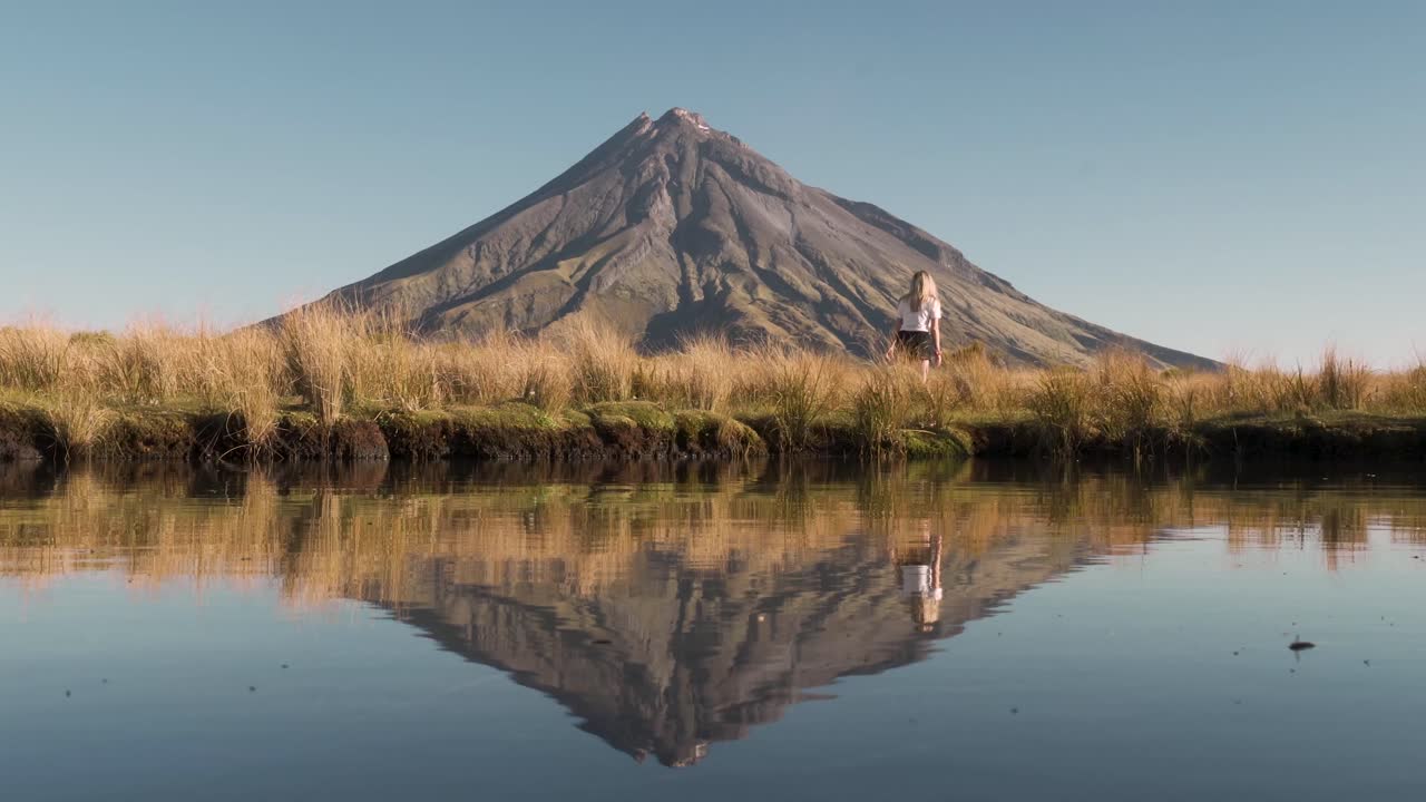 mujer rubia caminando a través de la hierba dorada frente al lago espejo con la montaña en el fondo durante la puesta de sol